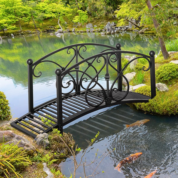Kleine dekorative Gartenbrücke aus schwarzem Metall mit geschwungenen Verzierungen über einem Teich mit Fischen in einem grünen Garten.