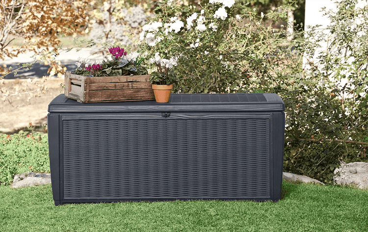 Hell gestalteter Garten mit anthrazitfarbener Gartenbox in Rattan-Optik, dekoriert mit Holzkiste und Blumen auf dem Deckel.