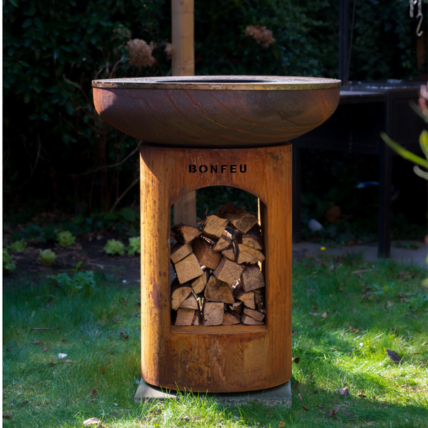 BonFeu Feuerschale und Grill aus Cortenstahl mit Holzaufbewahrung in einem Garten.