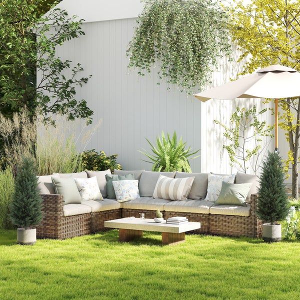 Garten-Ecksofa aus Geflecht mit hellen Polstern und einem niedrigen Couchtisch auf einem Rasen in einem hellen, organisierten Garten.