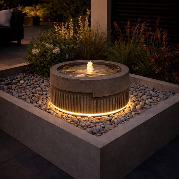 Runder Gartenbrunnen aus Stein mit Beleuchtung in einem Kiesbett und einer rechteckigen Betoneinfassung.