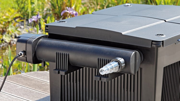 Schwarzes Teichfiltersystem mit integriertem UVC-Klärer auf einem Holzsteg neben einem bepflanzten Gartenteich.
