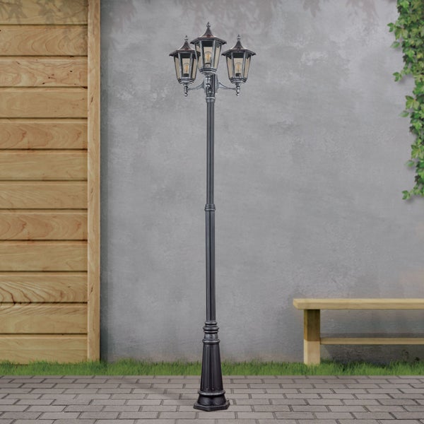 Dreiflammige Außenstehleuchte im Kandelaber-Stil auf einer gepflasterten Terrasse vor einer Wand in Steinoptik und einem Holzzaun.