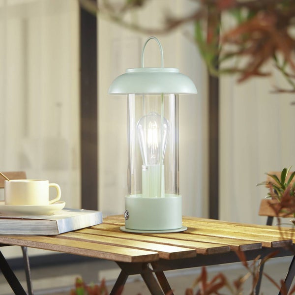 Hellgrüne tragbare LED-Tischleuchte im Laternen-Design auf einem hölzernen Gartentisch neben einer Tasse.