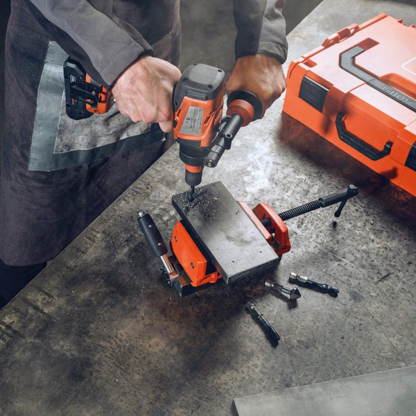 Bohren mit einer Akku-Magnetbohrmaschine in ein Werkstück, befestigt in einem Schraubstock. Eine Werkzeugbox befindet sich im Hintergrund.