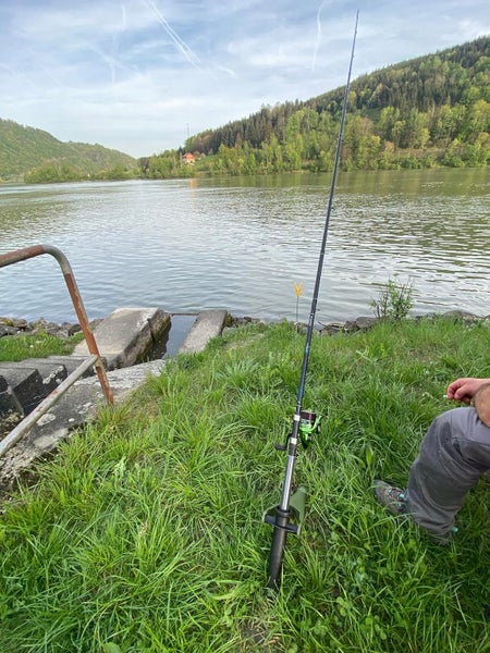 Angelrute mit Angelrolle auf einem Rutenhalter am Flussufer im Gras vor bewaldeten Hügeln.