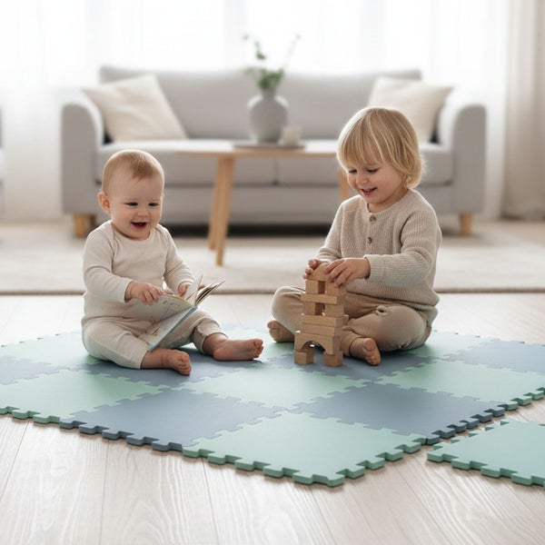 Kleinkinder spielen auf einer Schaumstoff-Puzzlematte im hellen Wohnzimmer. Ein Kind baut mit Holzklötzen, das andere betrachtet ein Buch.
