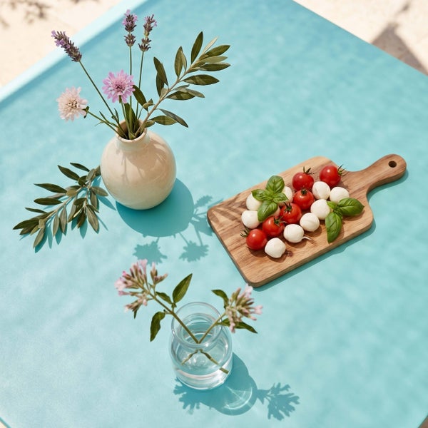 Blauer Gartentisch mit Keramikvase, Glasgefäß mit Blumen und Holzbrett mit Tomaten und Mozzarella in hellem Sonnenlicht.