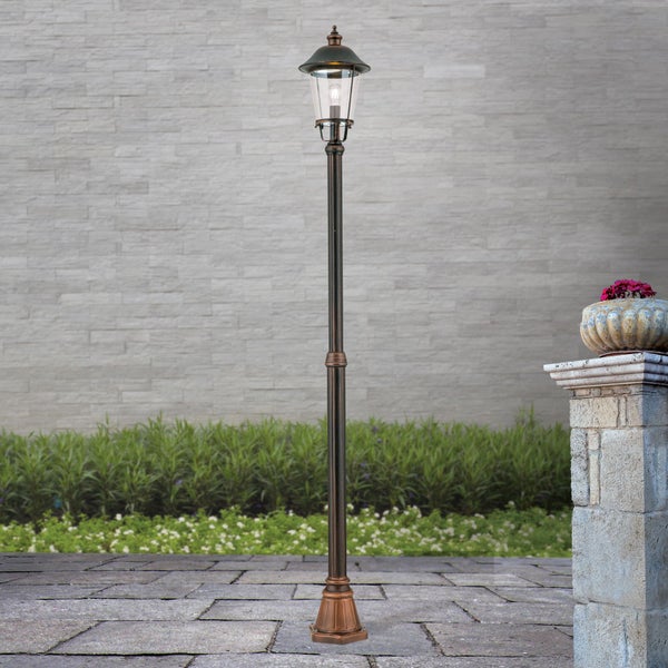 Klassische Außen-Mastleuchte mit bronzefarbenem Metallgestell und Glaslaterne auf einer Steinterrasse in einem hellen Garten.