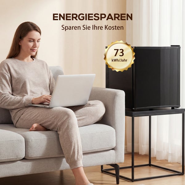 Frau auf Sofa mit Laptop neben schwarzem Minikühlschrank mit 73 Kilowattstunden pro Jahr Energieverbrauch in hellem Wohnzimmer.