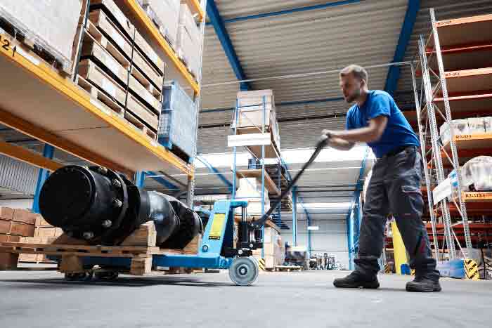 Ein Mitarbeiter bewegt mit einem Hubwagen eine schwere Last auf einer Palette in einem Lager mit Hochregalen.