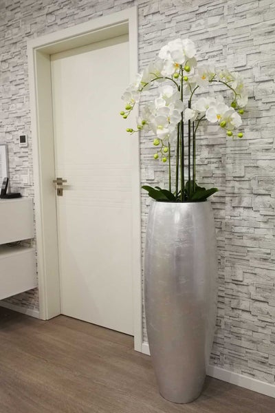 Große silberne Bodenvase mit weißen Orchideen neben einer weißen Innentür an einer Wand mit grauer Steinoptik-Verblendung.