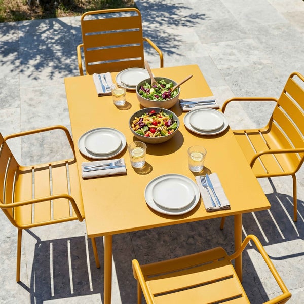 Gelber Garten-Esstisch aus Metall mit vier passenden Stühlen, gedeckt mit Geschirr und Salatschüsseln auf einer hellen Steinterrasse.