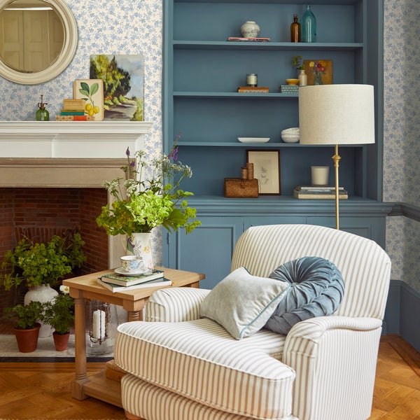 Wohnzimmer mit gestreiftem Sessel, blauem Einbauregal, Kamin und Tapete mit Blumenmuster im Landhausstil.