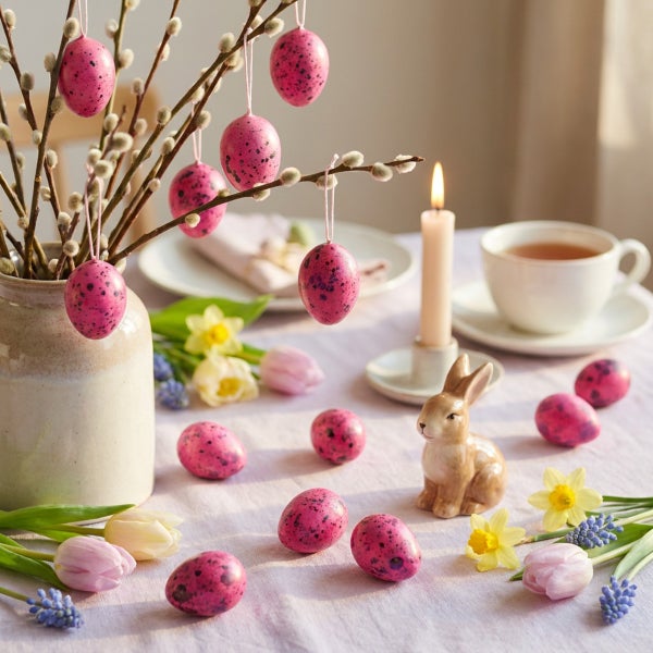 Osterdekoration auf einem gedeckten Tisch mit gesprenkelten Eiern, Weidenzweigen in einer Vase, einer Kerze, einem Osterhasen und Frühlingsblumen.