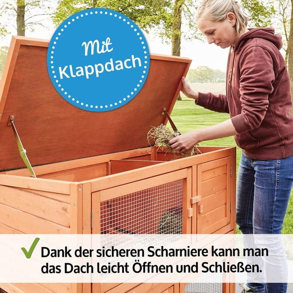 Eine Frau öffnet das Klappdach eines hölzernen Kleintierstalls mit sicherem Scharnier, um Stroh hineinzulegen.
