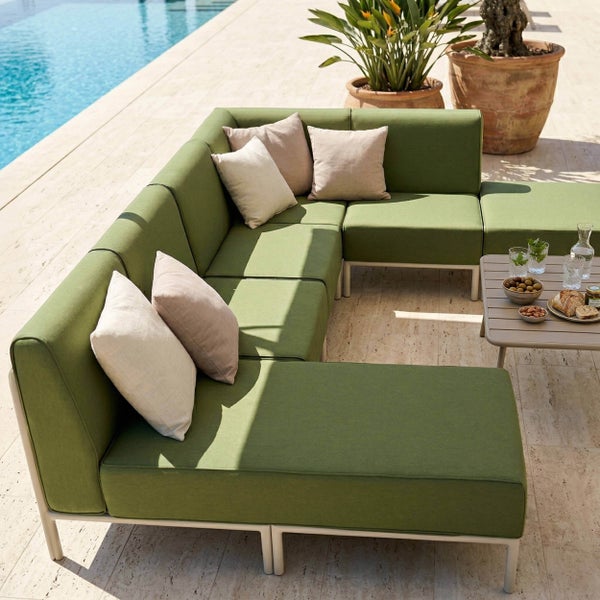 Modulares grünes Garten-Lounge-Sofa auf Steinterrasse am Pool mit beigen Kissen, Beistelltisch und Pflanzen in Terrakottatöpfen.