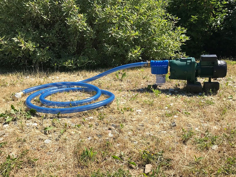 Grüne Gartenpumpe mit blauem Vorfilter und spiralförmigem blauem Saugschlauch auf einer Rasenfläche vor einem Gebüsch.