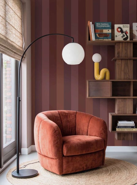 Ein wohnlicher Bereich mit einem samtigen Sessel vor einer gestreiften Wand, ergänzt durch eine Bogenlampe und ein modernes Regal.