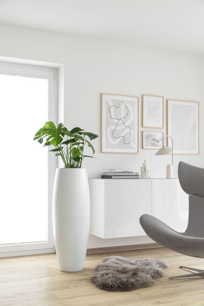 Hohe weiße Bodenvase mit Monstera-Pflanze in einem hellen, minimalistischen Wohnzimmer mit weißem Sideboard und Bilderwand.