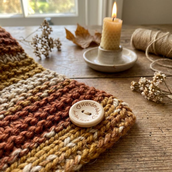 Handgestrickter Schal aus Wolle mit Holzknopf und der Aufschrift Handmade with love, dekoriert mit einer Kerze und herbstlichen Elementen.