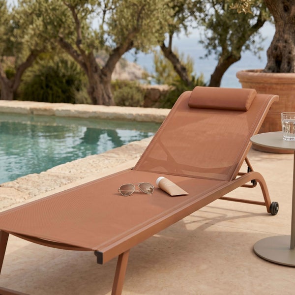 Sonnenliege mit Kopfstütze und Rollen an einem Pool, mit Beistelltisch, Sonnenbrille und Sonnencreme in einer hellen Außenanlage mit Bäumen und Meer.