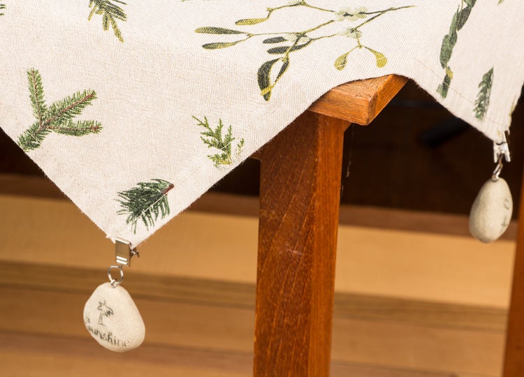 Tischdecke mit weihnachtlichem Zweigemuster und Tischdeckengewichten aus Stein an einem Holztisch