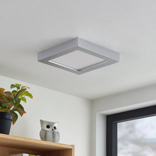 Moderne quadratische LED-Deckenleuchte mit silberfarbenem Rahmen in einem hellen Raum über einem Holzregal mit Pflanze und Eulenfigur.