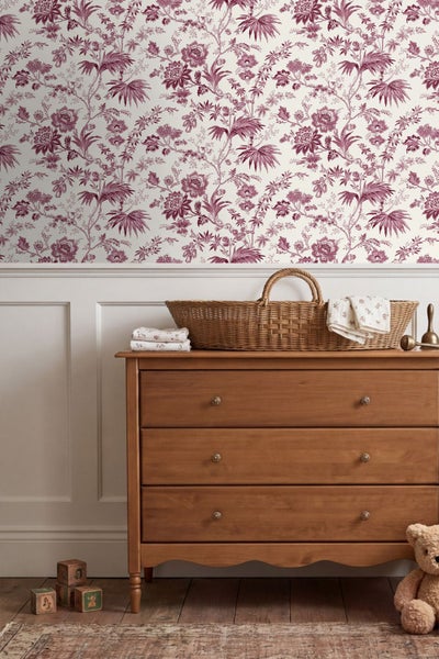 Helles Kinderzimmer mit floral gemusterter Tapete, weißer Wandvertäfelung, einer Holzkommode mit geflochtenem Korb und Spielzeug auf Dielenboden.