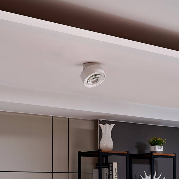 Weißer, moderner Aufbaustrahler an einer weißen Decke montiert, im Hintergrund ein Regal mit Dekorationsgegenständen.