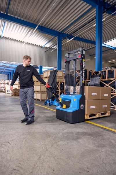 Ein Mitarbeiter bedient einen elektrischen HanseLifter Hochhubwagen in einem hellen, organisierten Lager, beladen mit Kartons auf einer Holzpalette.