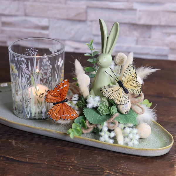 Osterdekoration mit Kerze, Hasenfigur und Schmetterlingen auf einem Teller