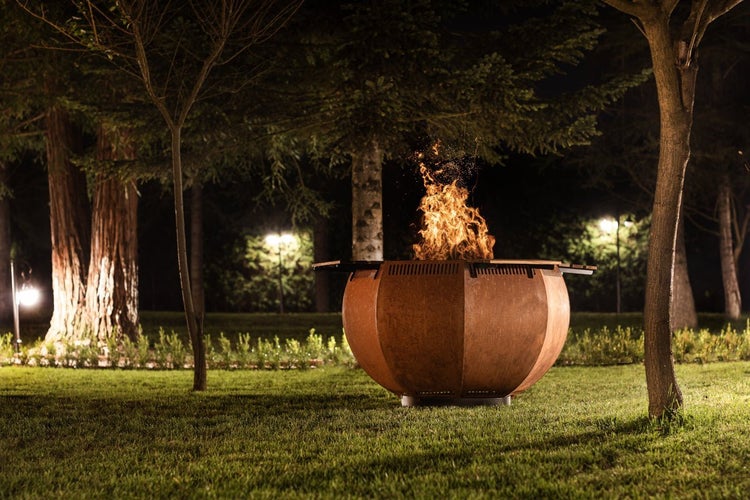 Eine brennende Feuerschale aus Cortenstahl mit Grillfunktion steht nachts in einem Garten.