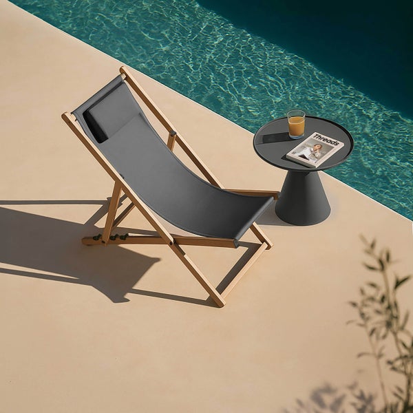 Klappbarer Liegestuhl aus Holz mit anthrazitfarbenem Stoff und Kopfkissen neben einem runden Beistelltisch an einem Pool.