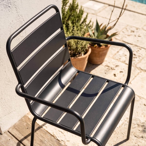 Moderner Gartenstuhl aus Metall mit Armlehnen und Lattendesign auf einer hellen Terrasse mit Pflanzen.