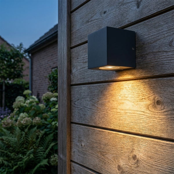 Schwarze würfelförmige Außenwandleuchte an einer Holzwand mit warmweißem Lichtkegel nach unten im Garten.