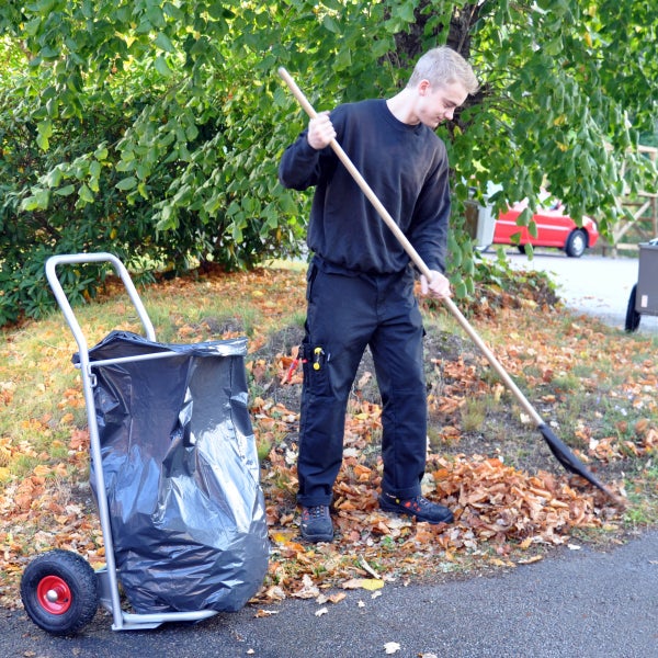 Person fegt trockenes Laub im Garten neben einem fahrbaren Müllsackständer für Gartenabfälle.