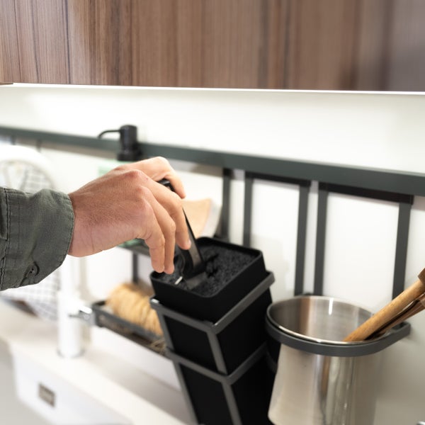 Hand greift nach einem Utensilienhalter an einer Küchenreling unter einem Wandschrank in einer organisierten Küche.