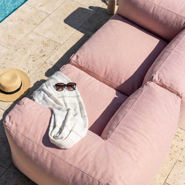 Rosafarbene Gartenlounge am Pool mit Strohhut, Sonnenbrille und gestreiftem Handtuch auf hellen Steinplatten.