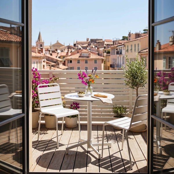 Moderner Balkon mit Holzdiele, zwei weißen Metallstühlen und einem runden Bistrotisch, umgeben von Pflanzen mit Blick auf eine Stadt.