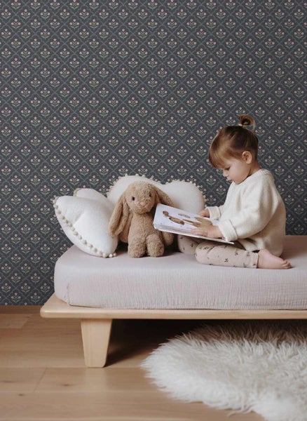 Kinderzimmer mit floral gemusterter Tapete, ein Kind liest ein Buch auf einem niedrigen Bett neben einem Stoffhasen und Kissen.