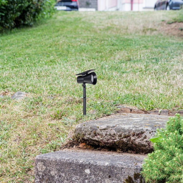 Solarbetriebener Gartenstrahler in Schwarz mit Erdspieß, platziert auf einer Rasenfläche neben Treppenstufen aus Stein.