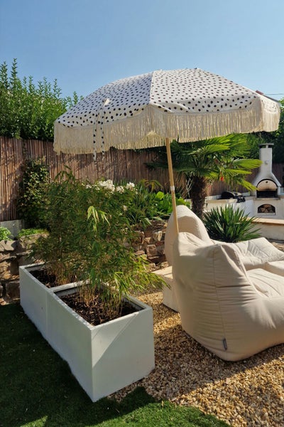 Moderner Garten mit gemustertem Sonnenschirm, beigen Sitzsäcken und weißen Pflanzgefäßen mit Bambus auf Kies.