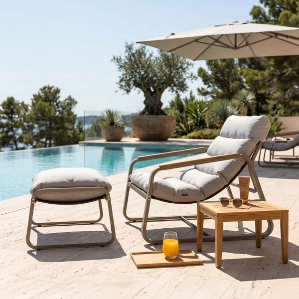 Moderner Relaxsessel mit Fußhocker und Holz-Beistelltisch auf einer hellen Terrasse am Pool, graue Stoffpolsterung und Metallgestell.