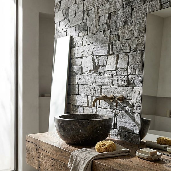Badezimmer mit rundem Naturstein Waschbecken auf einem massiven Holztisch vor einer rustikalen Steinwand mit Wandarmatur und Spiegel.