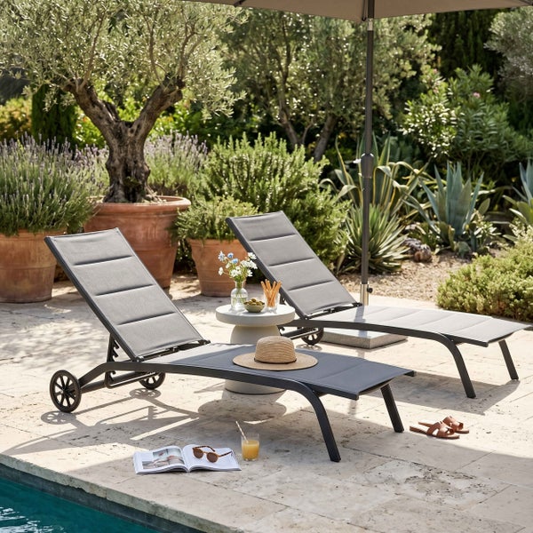 Zwei graue Sonnenliegen mit Rollen und verstellbarer Lehne auf einer Steinterrasse am Pool, umgeben von einem Garten mit Olivenbäumen.
