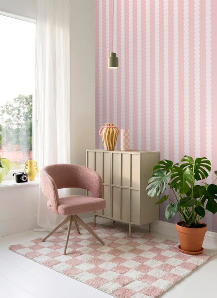 Helles Zimmer mit rosa-weiß gemusterter Vliestapete, rosa Polstersessel, beigem Sideboard, kariertem Teppich und einer Monstera-Pflanze.
