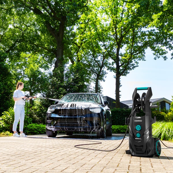 Frau reinigt ein schwarzes Auto mit einem VONROC Hochdruckreiniger auf einer gepflasterten Auffahrt vor grünen Bäumen.