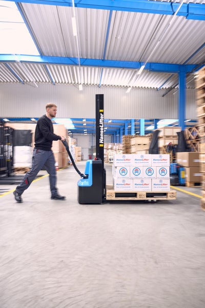 Ein Mitarbeiter bedient einen HanseLifter Elektro-Hochhubwagen in einem hellen Lagerhaus, um eine Palette mit Kartons zu transportieren.