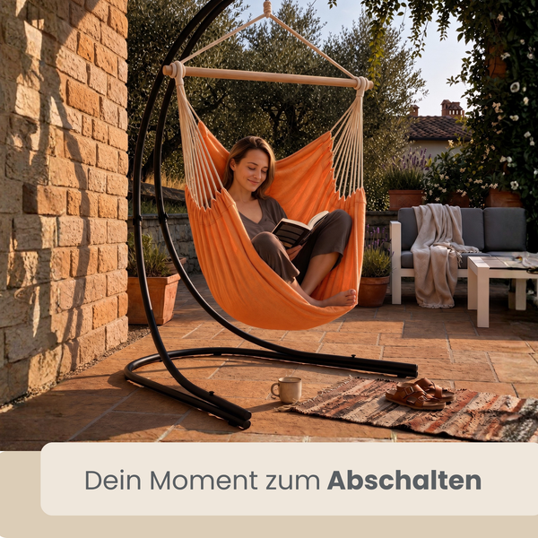 Frau sitzt in einem Hängesessel mit Gestell auf einer Terrasse und liest ein Buch.